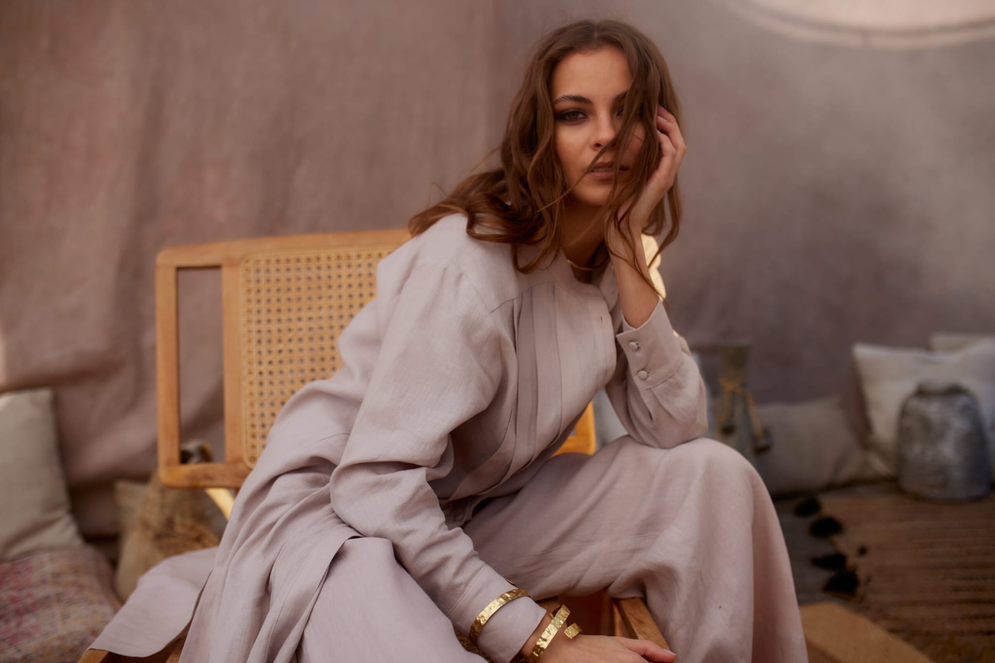 Model in a powder-beige linen dress, seated on a rattan chair in a sunlit interior